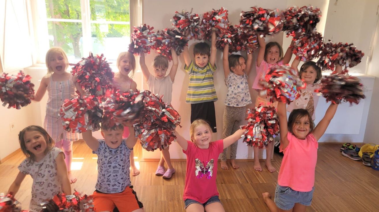 A group of young children, all holding red and silver pom-poms, are smiling and posing for a photo. They are standing in a line, with a white wall behind them.