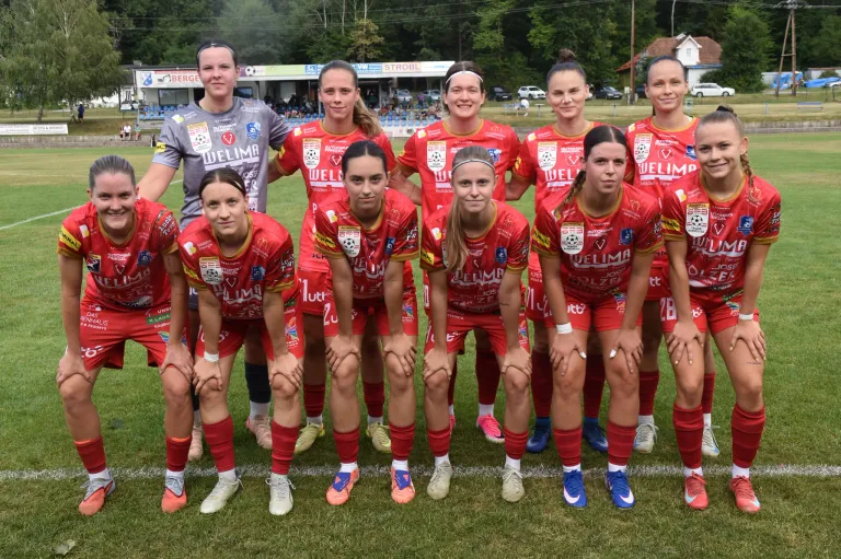 Eine Gruppe von Fußballerinnen in roten Trikots posiert für ein Teamfoto auf einem Rasenfeld.