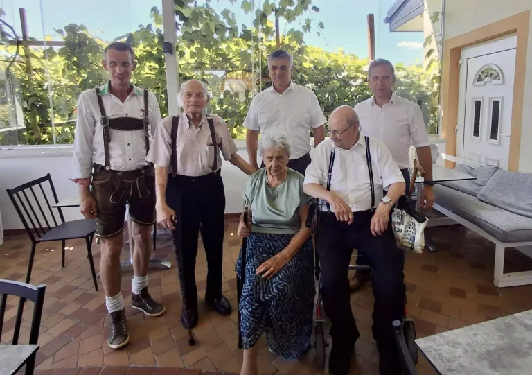 A group of elderly individuals, including a woman in a wheelchair, are standing together in a garden. They are all dressed in traditional clothing.