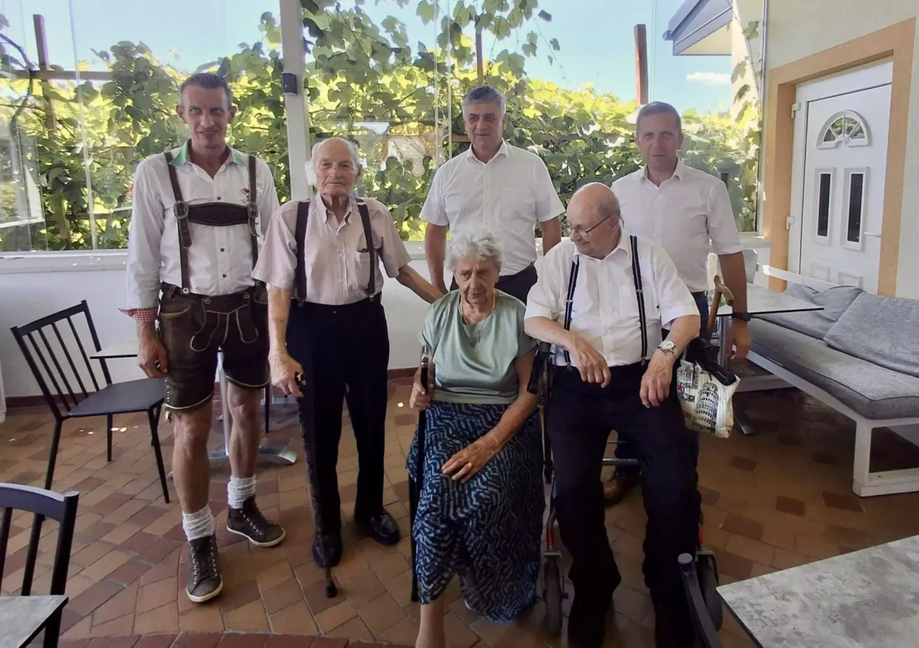 A group of elderly individuals, including a woman in a wheelchair, are standing together in a garden. They are all dressed in traditional clothing.