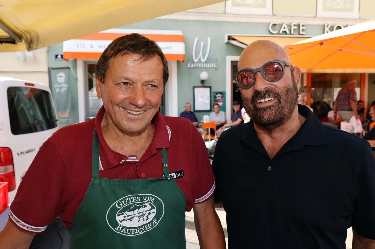Two men smiling in front of a cafe, one wearing a red shirt and a green apron with the text 'Gutes vom Bauernhof', the other in a black shirt and sunglasses.