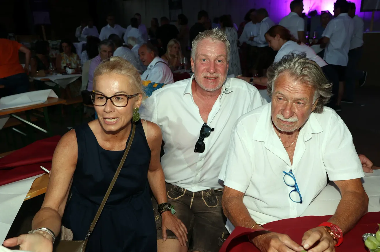 Three people are smiling and posing for a photo at a social event. The woman on the left wears glasses and a black sleeveless dress. The man in the middle has sunglasses on his shirt and is wearing a belt. The man on the right has a mustache and is wearing a white shirt.
