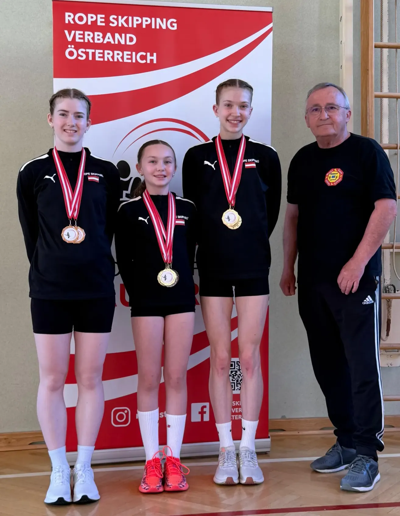 Drei junge Frauen und ein Mann stehen zusammen und tragen Medaillen. Die Frau links trägt eine Goldmedaille, die in der Mitte eine Silbermedaille und die rechts eine Goldmedaille. Der Mann trägt eine Brille und ein schwarzes T-Shirt.