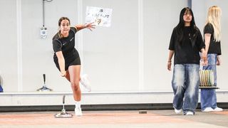 Eine Frau in einem schwarzen Outfit wirft einen Diskus in einer Turnhalle, während eine andere Frau zuschaut. Ein Schild mit der Aufschrift 'DIES IST KURZ' ist sichtbar.