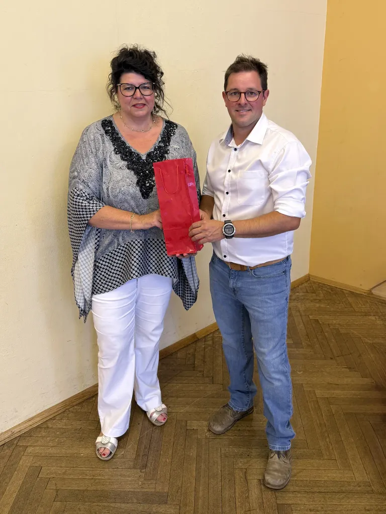 A woman in a patterned blouse and white pants holds a red gift bag while standing next to a man in a white shirt and jeans. They are both smiling and standing in a room with wooden flooring and a yellow wall.