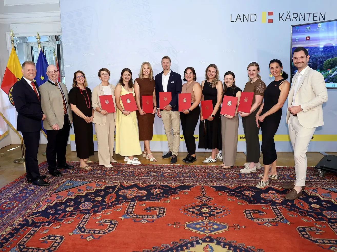 A group of people stand together holding red certificates in front of a backdrop with the word 'LAND'. They are dressed in formal attire and appear to be posing for a photo.