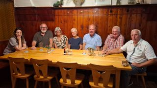 Fünf Personen sitzen um einen Holztisch und lächeln für ein Foto. Der Tisch hat Gläser, Blumen und Gewürze. Dahinter eine Holzwand mit gerahmten Bildern und Pflanzen.