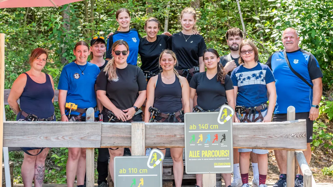 Eine Gruppe junger Menschen in passenden schwarzen Shirts und Gurten steht hinter einem Holzzaun und lächelt für ein Foto. Ein Schild vor ihnen zeigt 'ab 140 cm' und 'ALLE PARCOUR'.