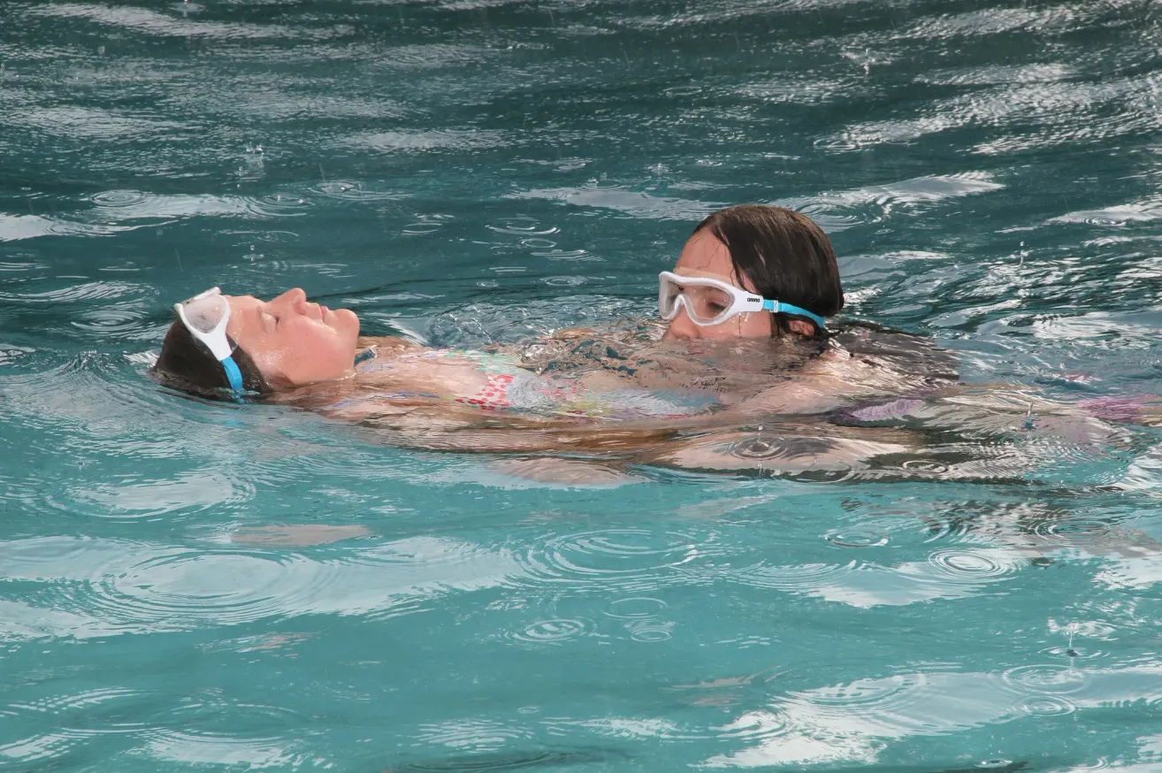 Two young girls wearing goggles are swimming in a pool, one lying on her back and the other on her stomach.