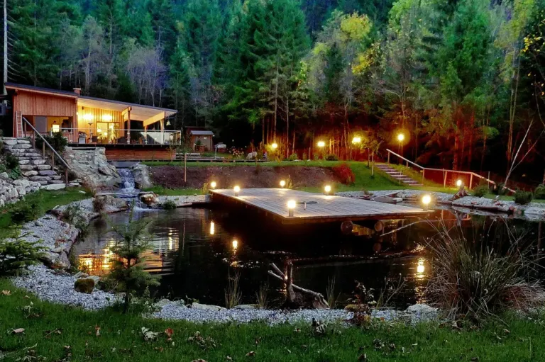 A serene pond with a wooden dock illuminated by lights, surrounded by trees and a small waterfall. A house with a balcony is visible in the background.