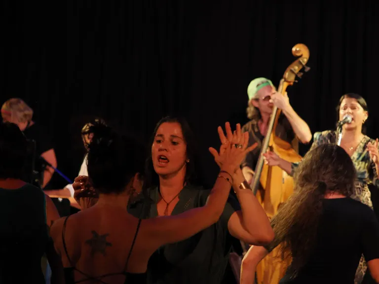 A woman with a tattoo on her back is singing and dancing with others on stage. Behind her, a man plays a cello.