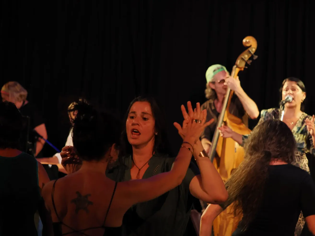 A woman with a tattoo on her back is singing and dancing with others on stage. Behind her, a man plays a cello.