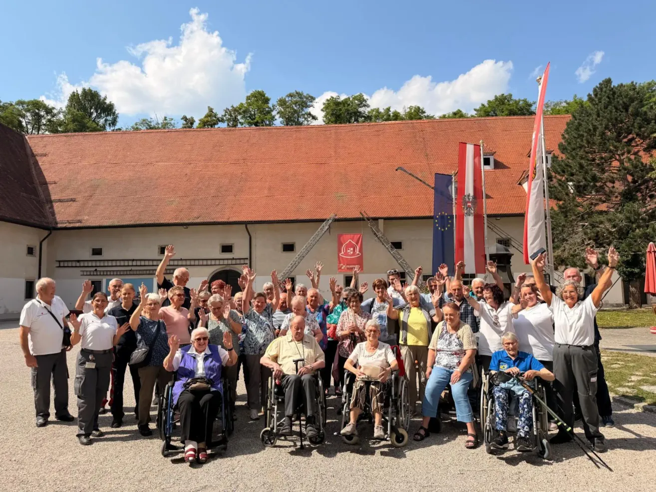 A group of elderly people in wheelchairs and standing pose for a photo in front of a building with flags.