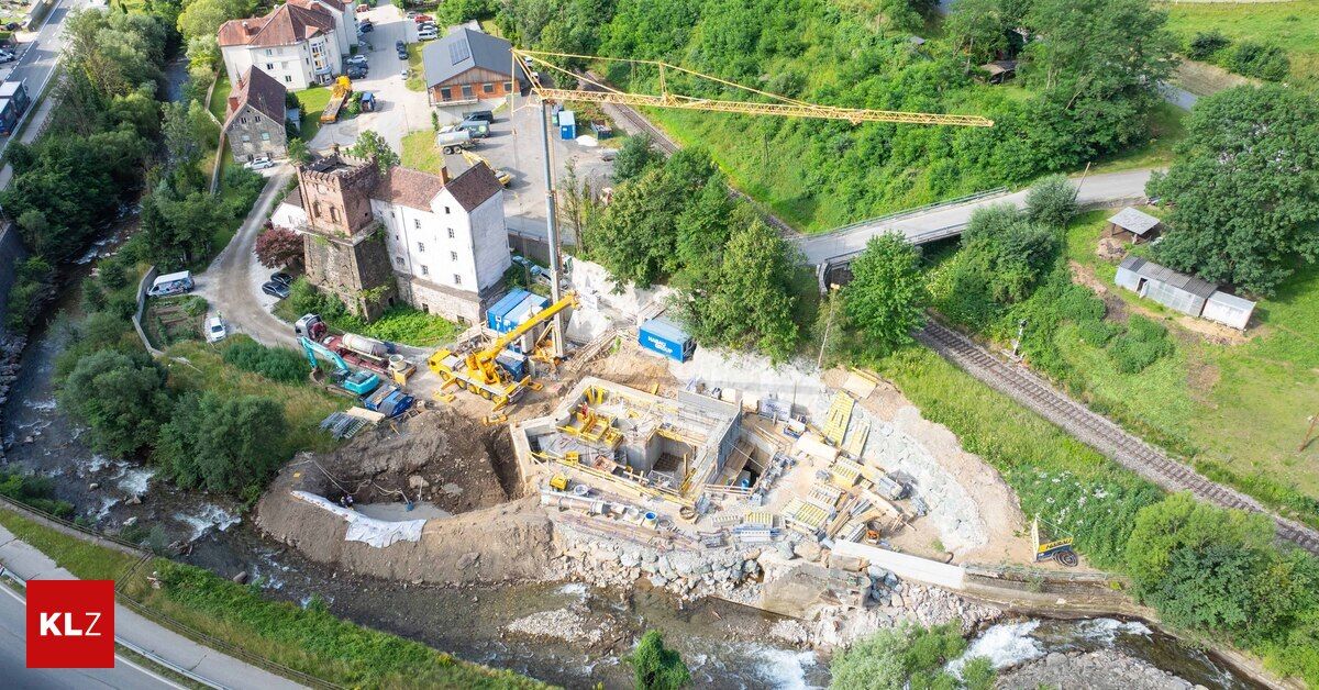 Eine Luftaufnahme einer Baustelle mit einem Kran, Maschinen und Arbeitern. Die Baustelle ist von einem Fluss, Bäumen und Häusern umgeben.