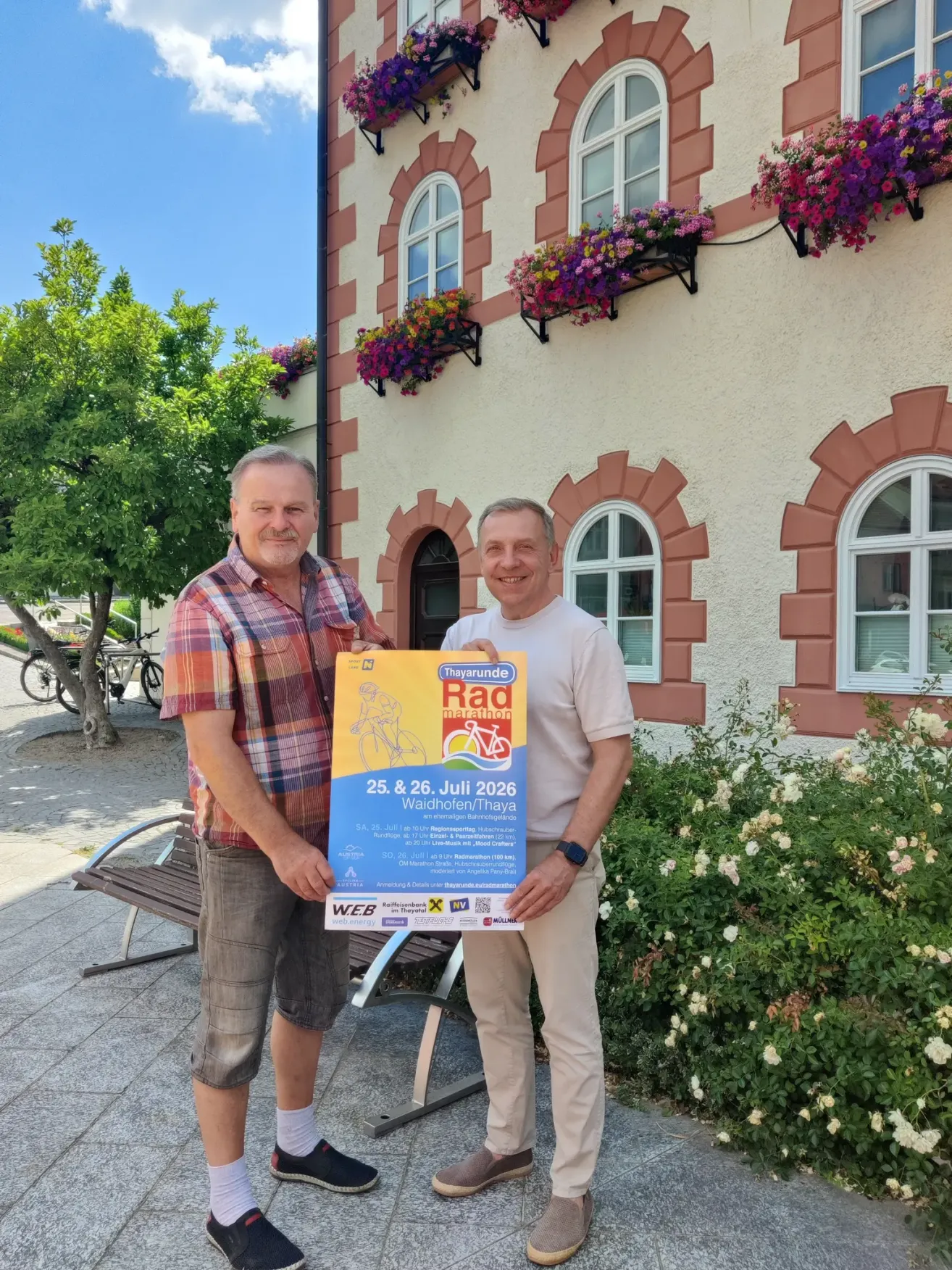 Two men stand in front of a building with a sign for the Radmarathon Thayrunde event on July 25 and 26, 2026.
