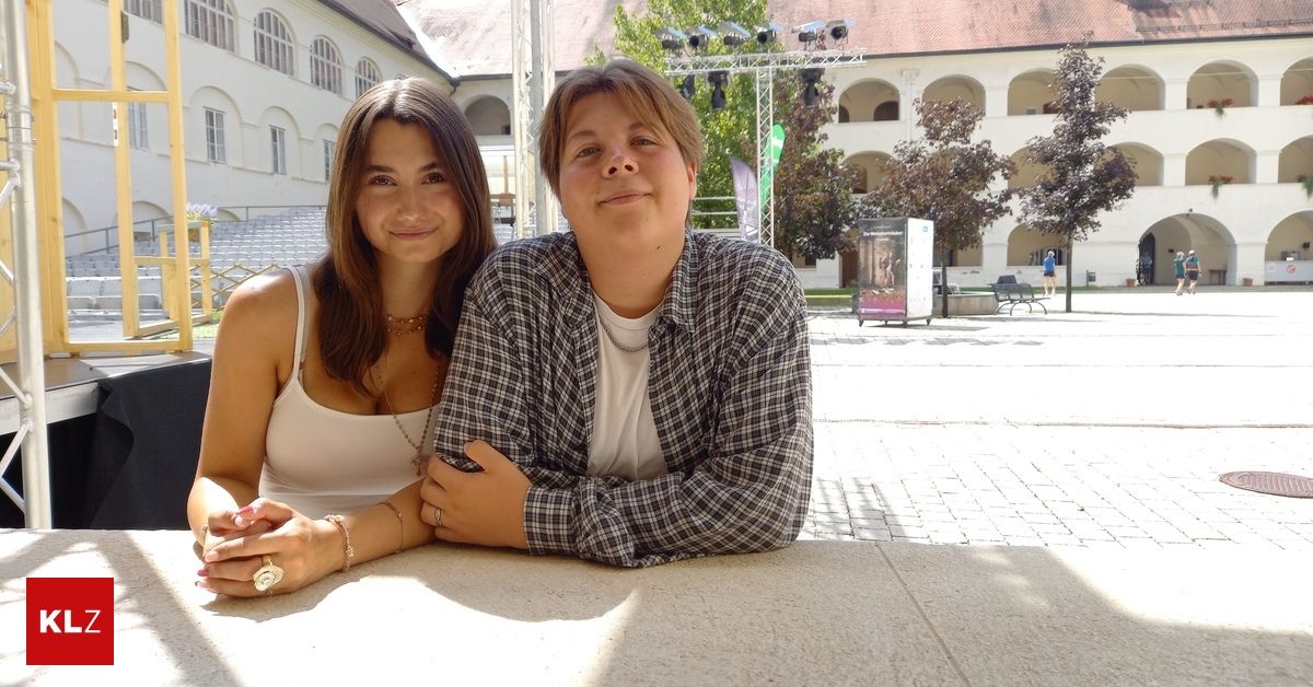 Zwei Personen sitzen auf einer Bank vor einem Gebäude mit einem Ziegelboden. Die Frau trägt eine Halskette und einen Ring. Der Mann trägt ein kariertes Hemd.