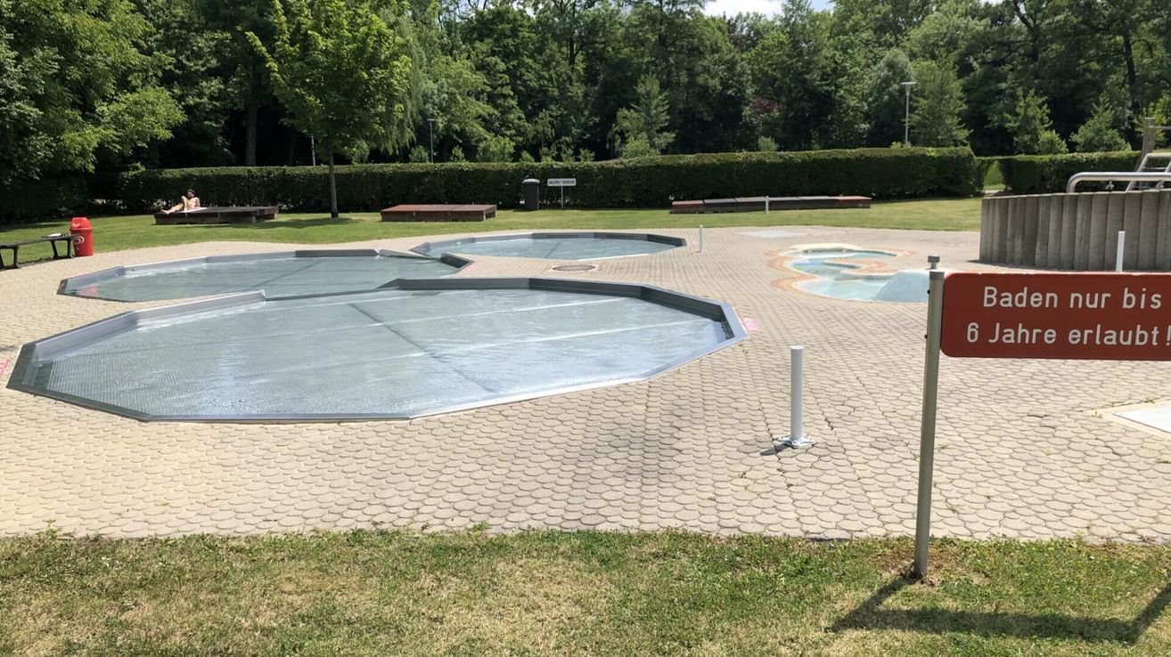 Three empty swimming pools covered with tarps are situated on a paved area, surrounded by a hedge and grass.