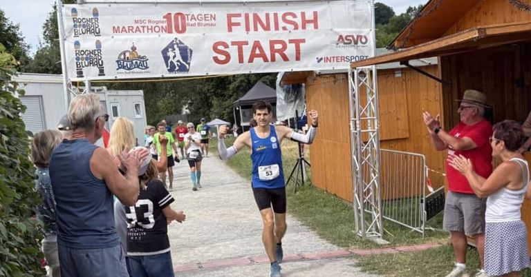 A marathon runner with the number 53 on his shirt crosses the finish line under a banner that reads 'Finish Start'. He is surrounded by other runners and spectators.