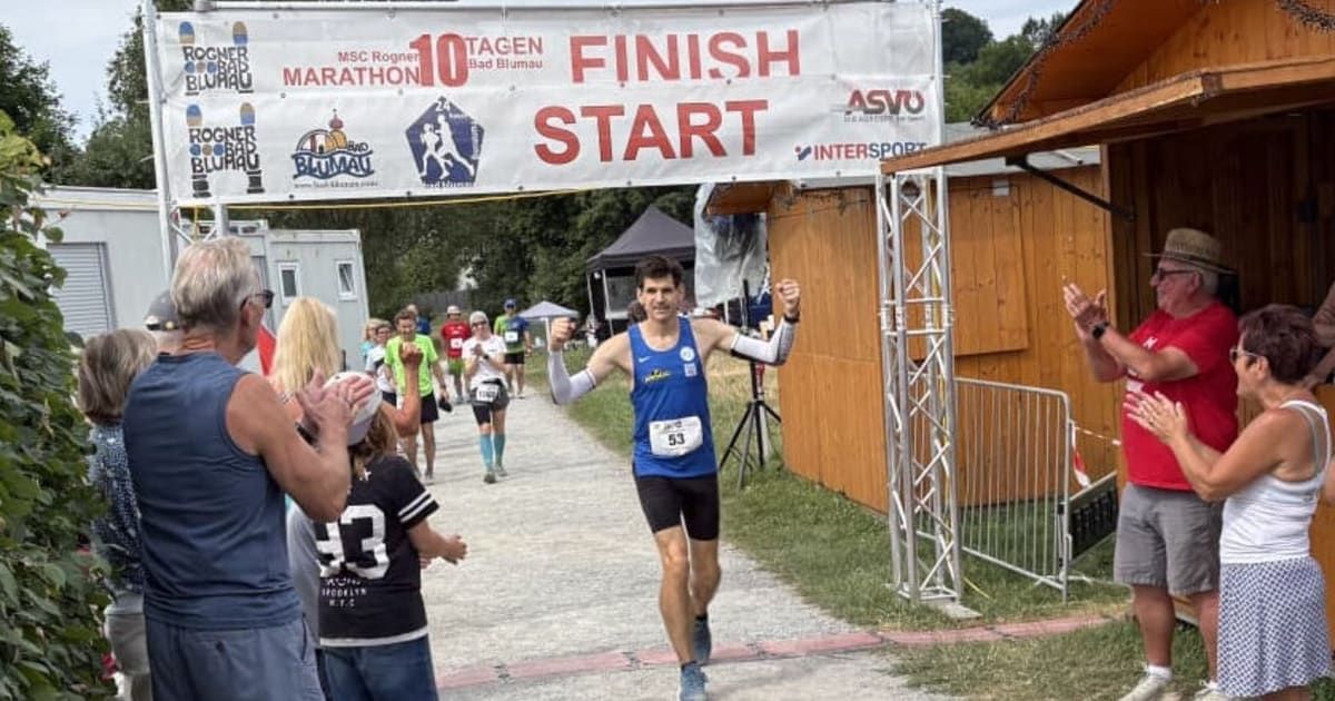 A marathon runner with the number 53 on his shirt crosses the finish line under a banner that reads 'Finish Start'. He is surrounded by other runners and spectators.