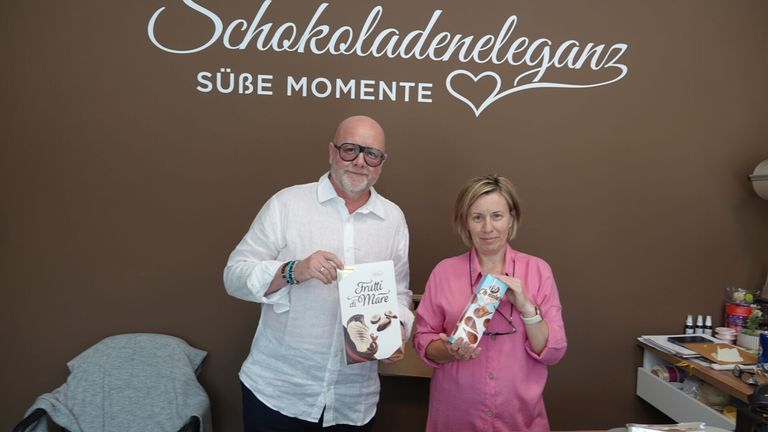 Ein Mann und eine Frau stehen vor einer Wand mit den Worten Schokoladenelegenz und Sube Momente und halten Schachteln mit Schokoladen.