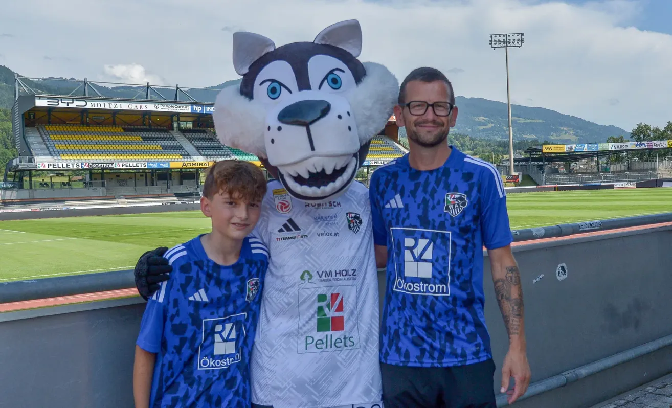 A mascot, a boy, and a man stand together in front of a stadium. The mascot is white with blue eyes, and the boy and man wear blue jerseys with logos.
