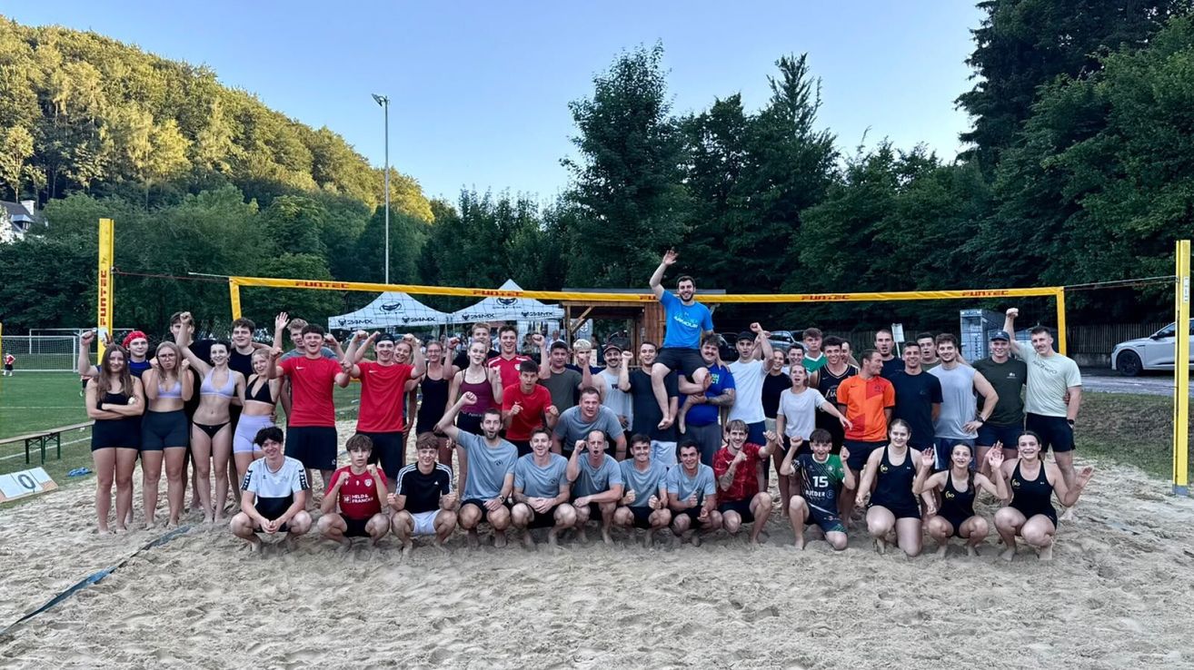 Eine Gruppe von Menschen in Sportkleidung posiert für ein Foto auf einem sandigen Strand mit einem Volleyballnetz im Hintergrund.