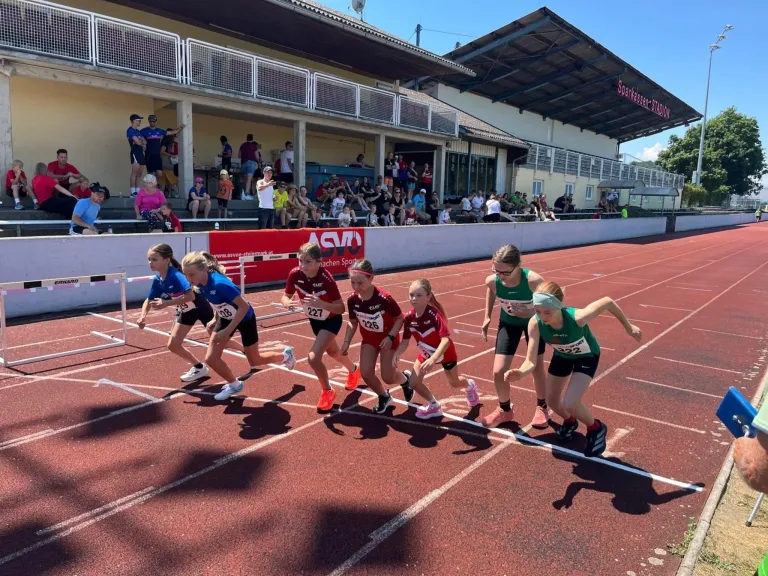 A group of young athletes compete in a track race at a stadium, with spectators watching from the stands.