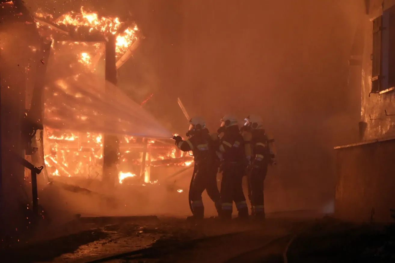 Drei Feuerwehrleute in voller Ausrüstung sprühen Wasser auf ein brennendes Gebäude. Das Feuer ist intensiv, und Rauch füllt die Luft.
