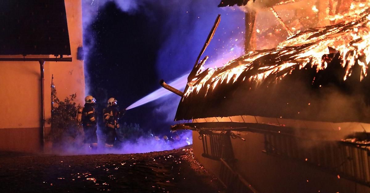 Feuerwehrleute in Schutzkleidung benutzen einen Schlauch, um ein brennendes Gebäude bei Nacht zu löschen. Das Dach steht in Flammen, und Rauch dringt heraus.