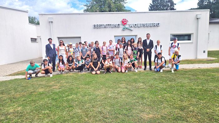 A group of children and adults pose for a photo in front of a white building with a sign reading 'Bestattung Wolfsberg'.