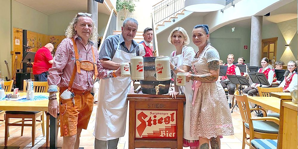 Four people in traditional German attire stand around a barrel labeled Stiegl Bier, holding mugs. Behind them, a band is seated with music stands.
