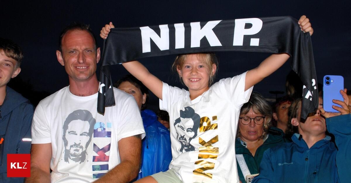 A young girl wearing a white t-shirt with a gold design and holding a black banner with white letters reading 'NIKP'. She is standing on someone's shoulders in a crowd.