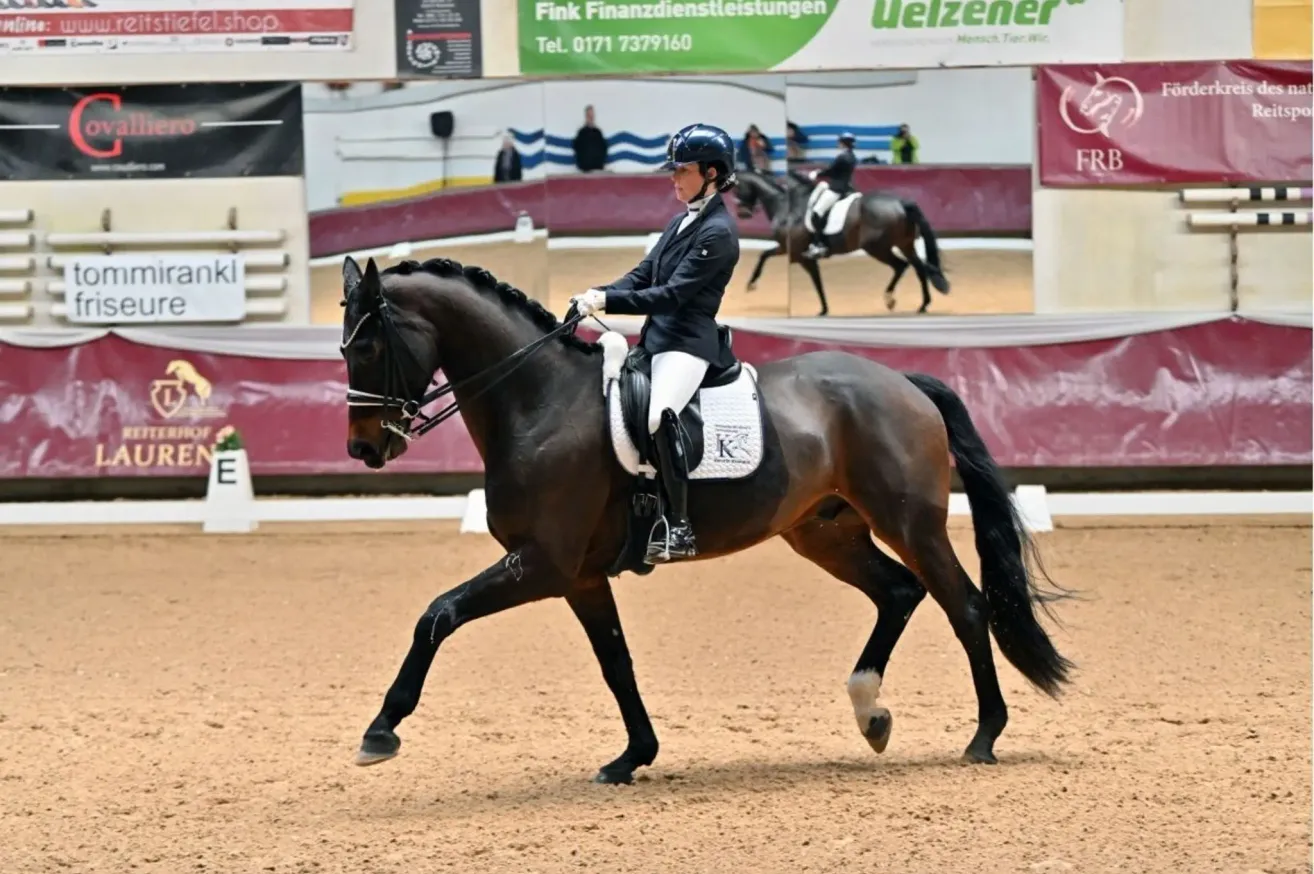 Eine Frau in einem schwarzen Reitanzug und Helm reitet ein braunes Pferd in einer überdachten Reithalle. Ein weiterer Reiter ist im Hintergrund.