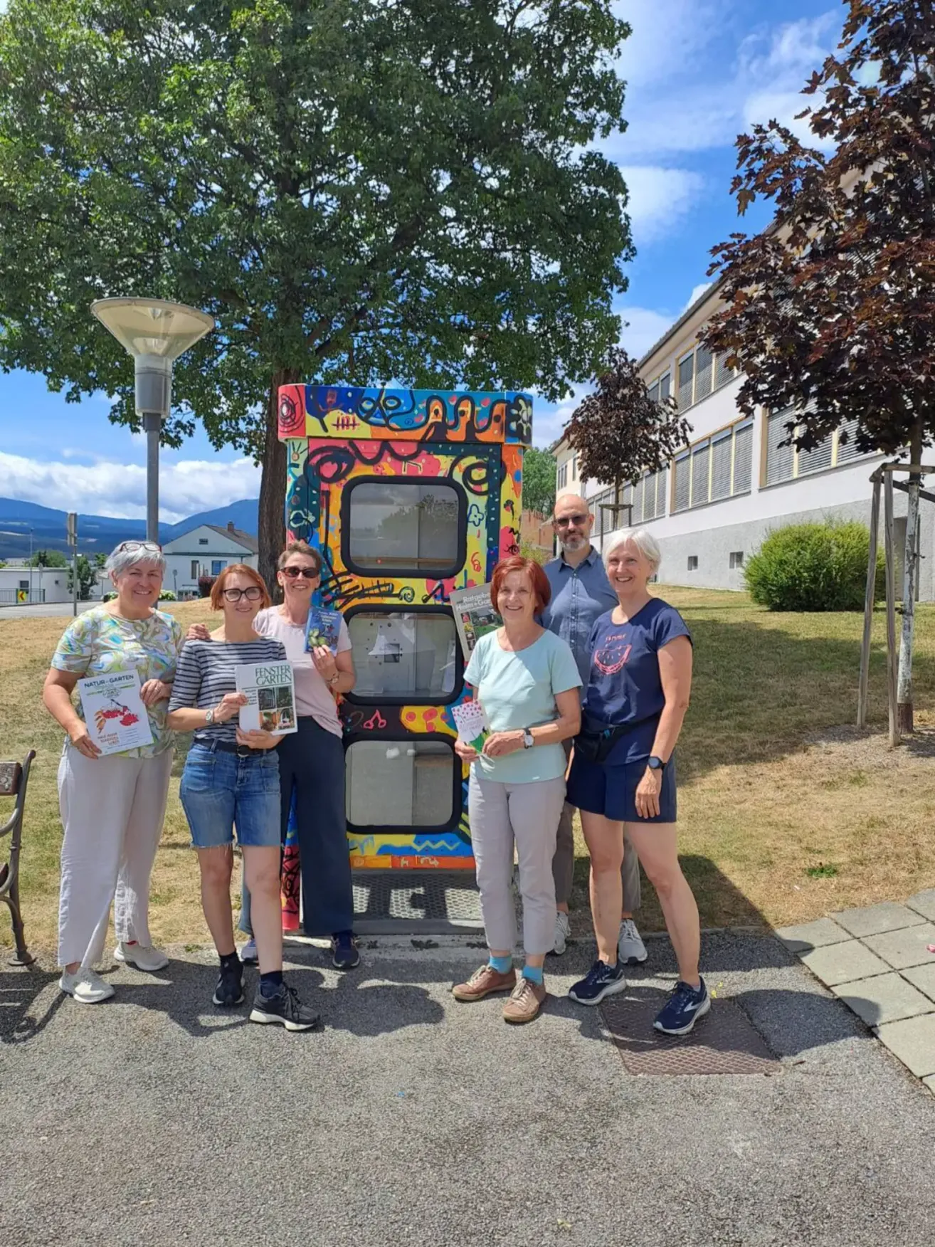 A group of people stand in front of a colorfully painted box, holding books and smiling for a photo. The setting is outdoors with trees and a building in the background.
