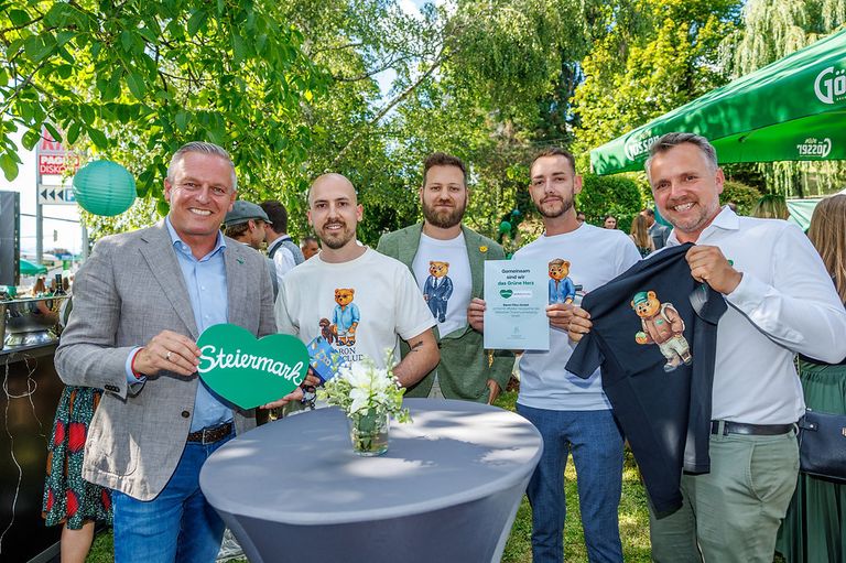 Five men stand in a park, smiling, with one holding a green heart sign that reads 'Steiermark'. Another holds a certificate, and a third holds a black shirt with a bear design. Behind them, there is a green tent and trees.