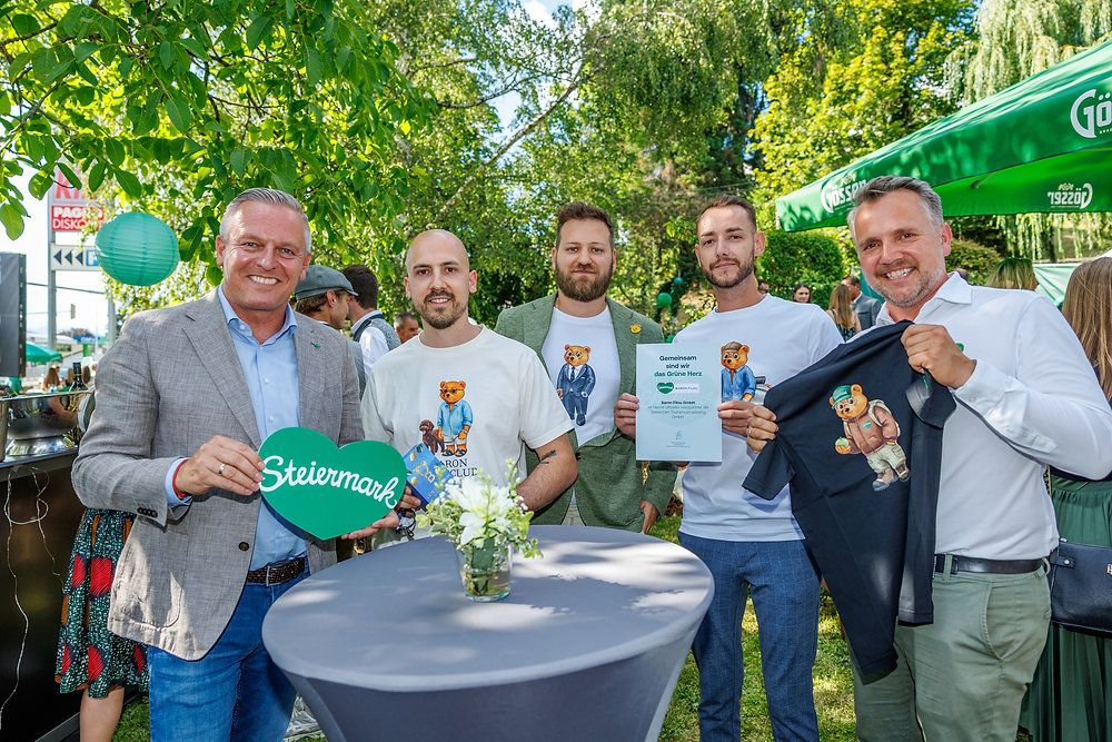 Five men stand in a park, smiling, with one holding a green heart sign that reads 'Steiermark'. Another holds a certificate, and a third holds a black shirt with a bear design. Behind them, there is a green tent and trees.