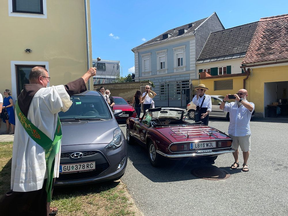 A group of people are gathered around vintage cars in a town square, with one man holding a bell and another taking a photo.