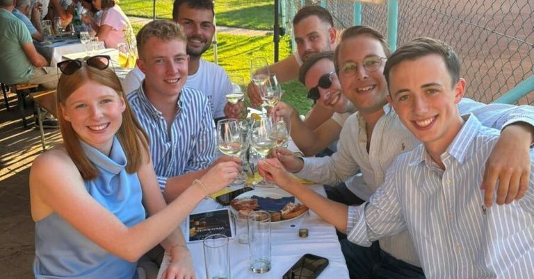 Eine Gruppe von Menschen sitzt an einem Tisch, lächelt und hält Weingläser. Sie stoßen mit ihren Getränken an. Es gibt Teller mit Essen, Gläser und ein Handy auf dem Tisch. Der Hintergrund zeigt einen Zaun und eine Rasenfläche.