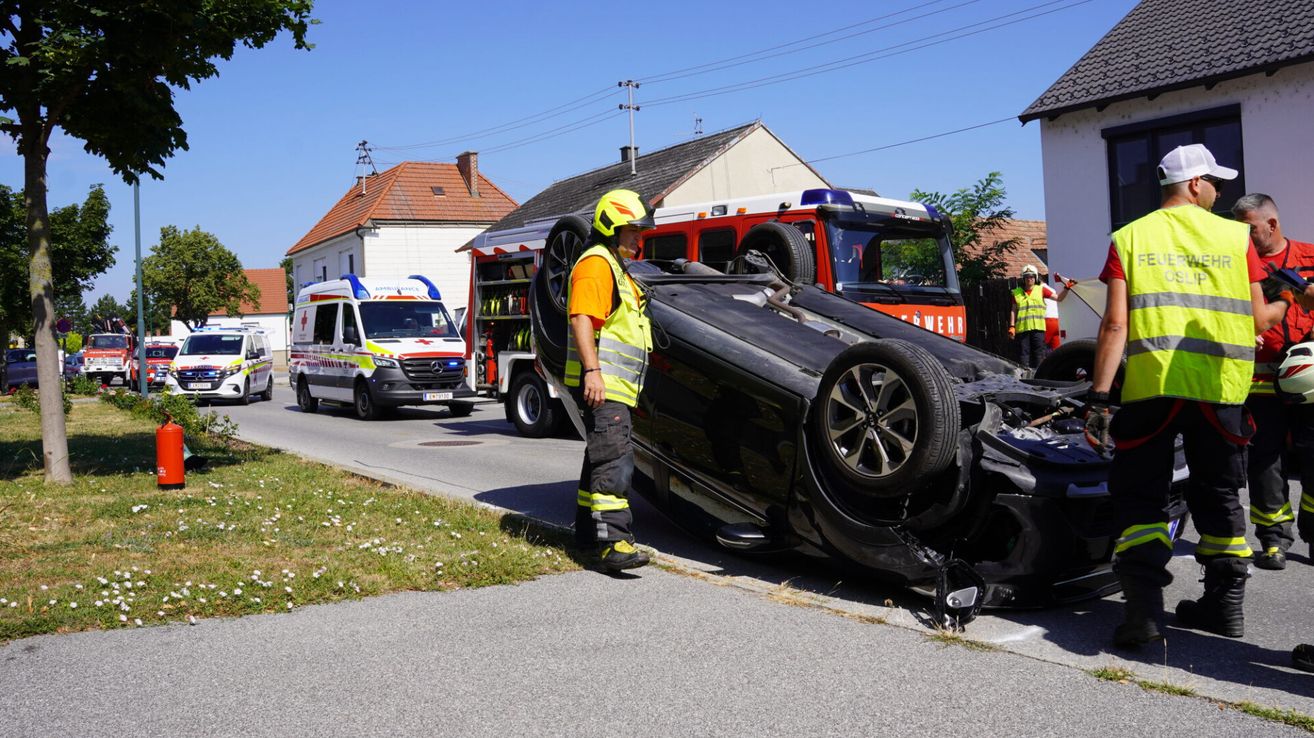 Ein umgekipptes Auto am Straßenrand mit nahegelegenen Rettungsfahrzeugen, eine Person in einer reflektierenden Weste steht neben dem Auto.