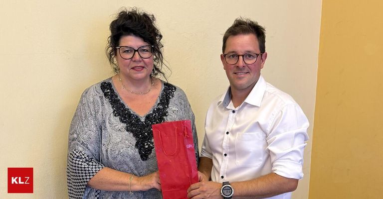 A woman wearing glasses and a necklace holds a red gift bag while standing next to a smiling man in a white shirt.