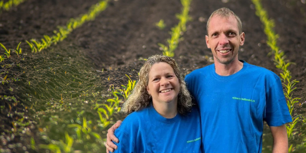 Ein Mann und eine Frau stehen in einem Feld, beide tragen blaue T-Shirts und lächeln für die Kamera.