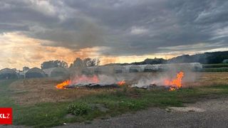A field with a fire burning in the middle, surrounded by green grass and a cloudy sky.