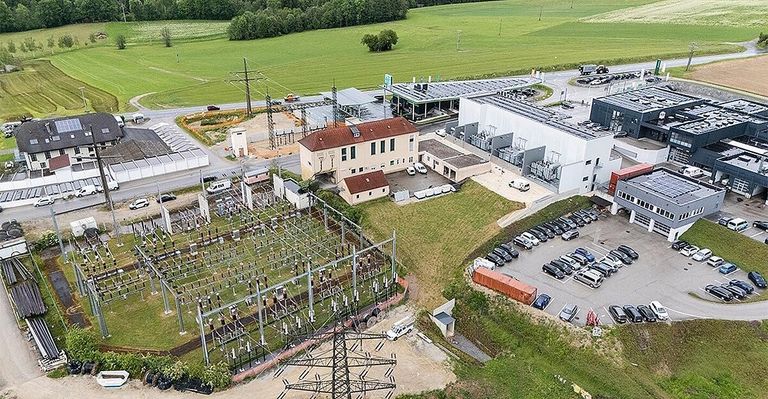 Eine Luftaufnahme eines Kraftwerks mit mehreren Gebäuden, einem gefüllten Parkplatz mit Autos und einem weitläufigen grünen Feld im Hintergrund.