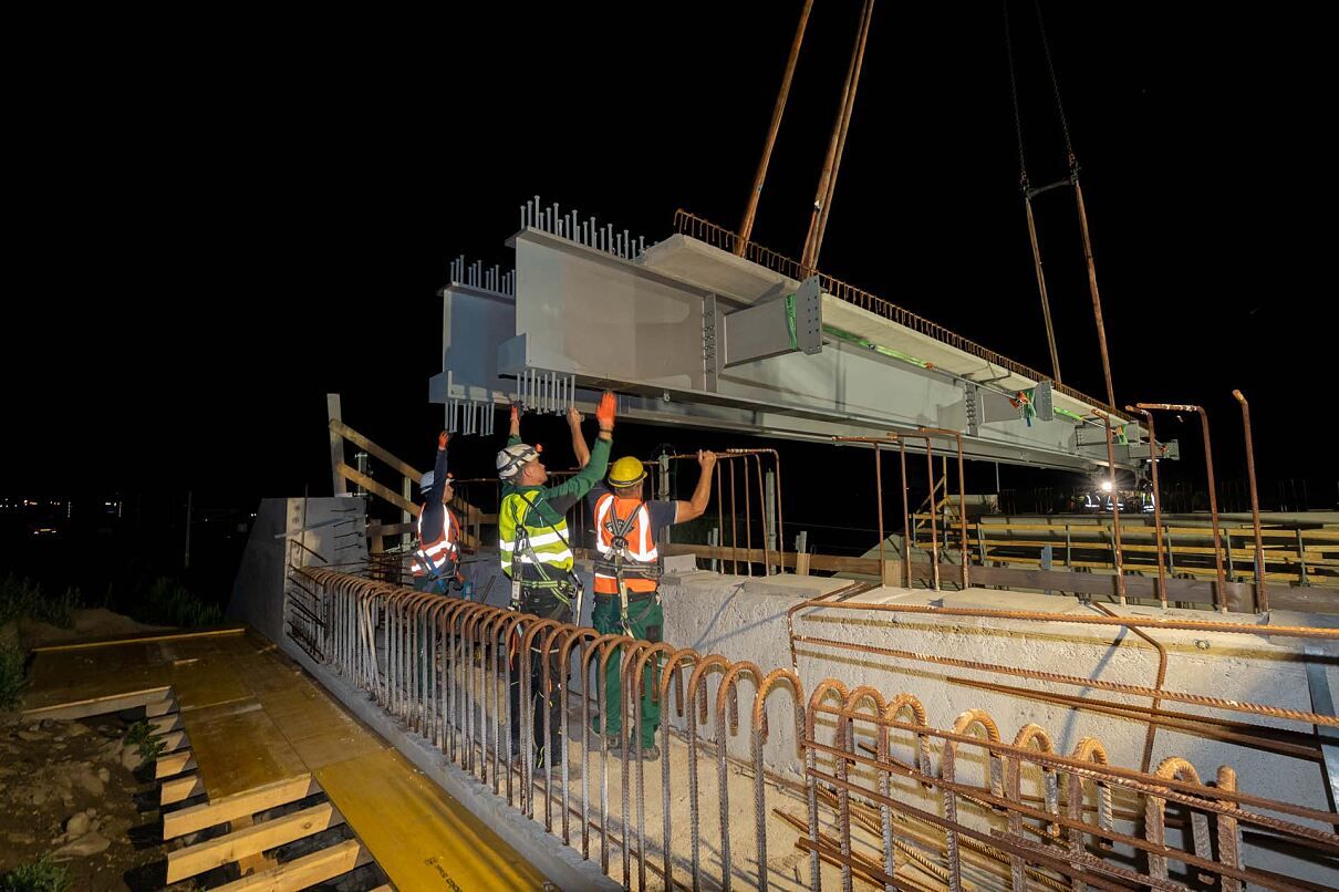 Vier Bauarbeiter führen einen großen Betonbalken bei Nacht auf eine Brücke. Sie tragen Sicherheitsausrüstung und Helme.