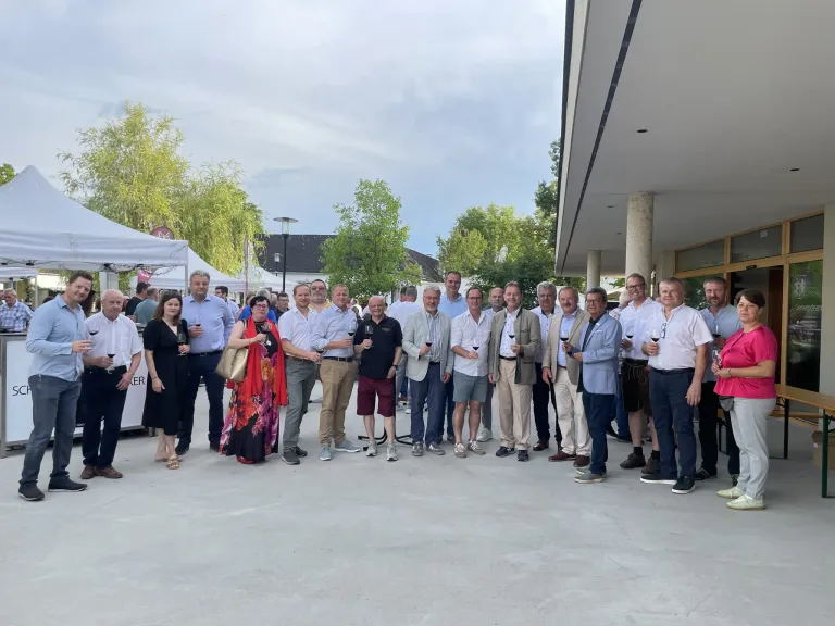 Eine Gruppe von Menschen in formeller Kleidung steht draußen und hält Weinglas in der Hand. Sie lächeln für ein Foto vor einem Gebäude mit Bäumen und Zelten im Hintergrund.
