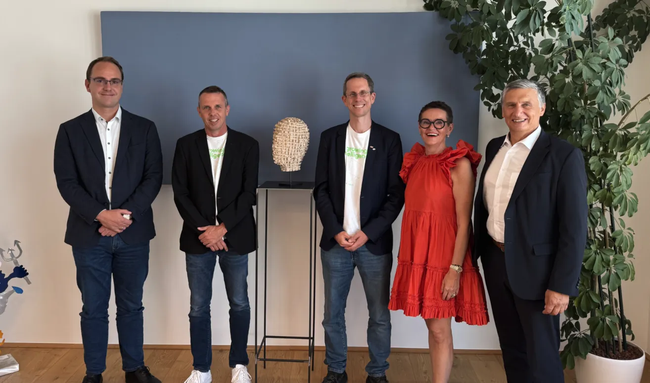 Fünf Personen stehen in einer Reihe und lächeln für ein Foto. Ein Mann auf der linken Seite trägt einen schwarzen Anzug mit einem grünen Logo, während der Mann neben ihm ein weißes T-Shirt mit einem Logo trägt. Die Frau in der Mitte trägt ein rotes Kleid. Eine Skulptur steht auf einem Ständer hinter ihnen.