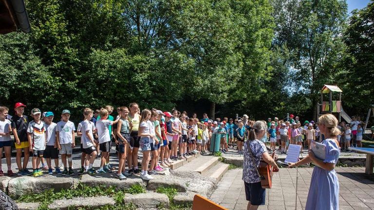 A group of children and adults stand in line outdoors, some holding musical instruments, with a woman playing a guitar and singing in the foreground.