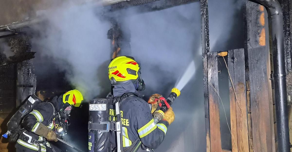 Ein Feuerwehrmann in voller Ausrüstung, einschließlich gelbem Helm und Maske, spritzt mit einem Schlauch Wasser auf ein brennendes Gebäude. Hinter ihm steht ein weiterer Feuerwehrmann, ebenfalls mit einem Schlauch ausgestattet.
