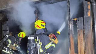 A firefighter in full gear, including a yellow helmet and mask, is using a hose to spray water on a burning building. Another firefighter is behind him, also equipped with a hose.