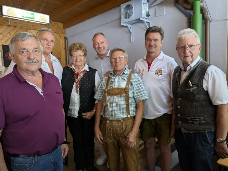 Eine Gruppe von Menschen steht zusammen in einem Raum, lächelt und posiert für ein Foto. Der Mann in der Mitte trägt ein kariertes Hemd und eine Hose mit Aufhängung. Hinter ihm trägt eine Frau einen Schal und eine Brille. Links trägt ein Mann ein lila Hemd. Rechts trägt ein Mann eine Weste.